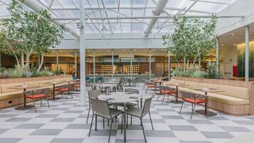 A spacious indoor area with multiple seating arrangements, including tables and sofas, surrounded by potted plants and a high ceiling with natural light.