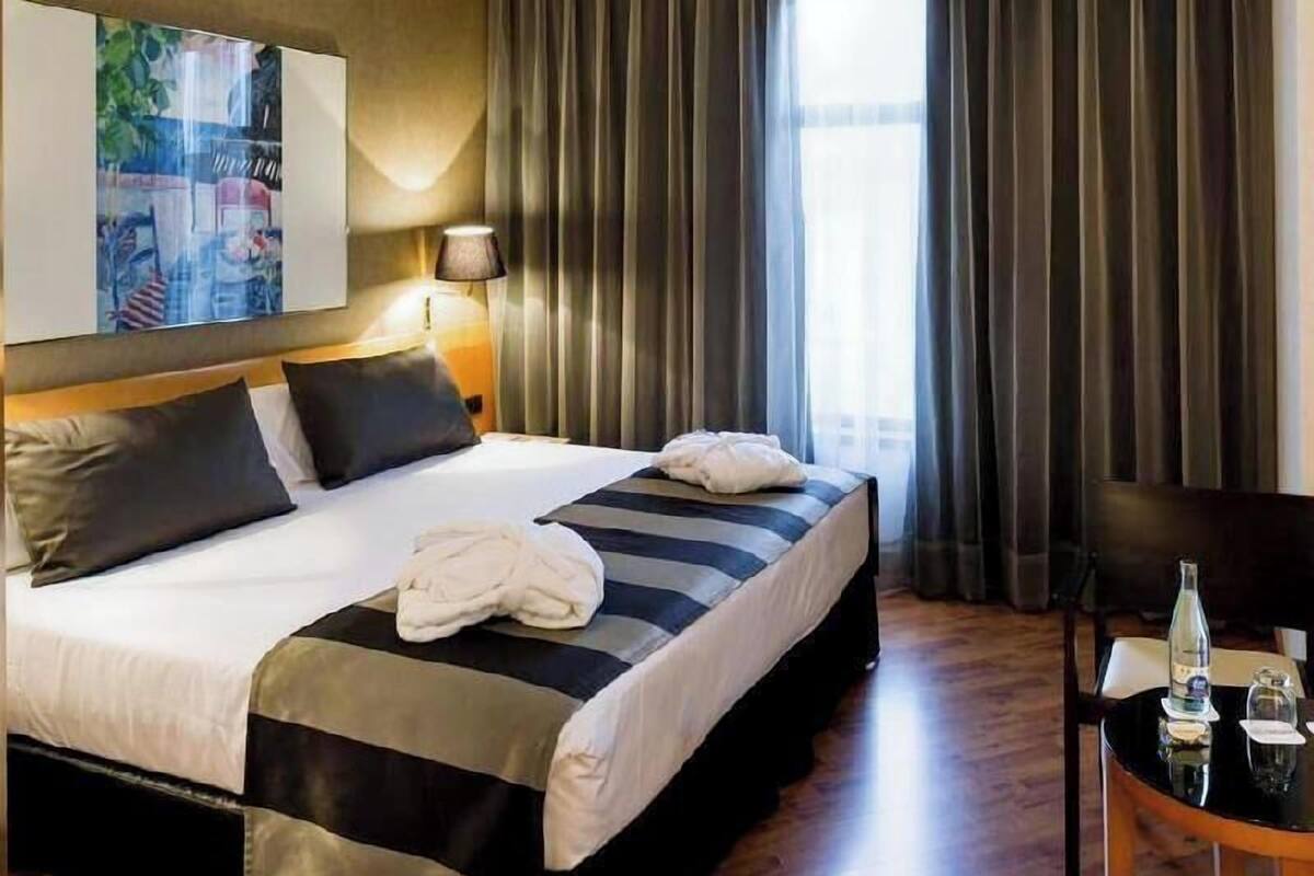 Une chambre d’hôtel avec un lit, une table de chevet sur laquelle se trouvent une bouteille et des verres, une lampe et un tableau accroché au mur.