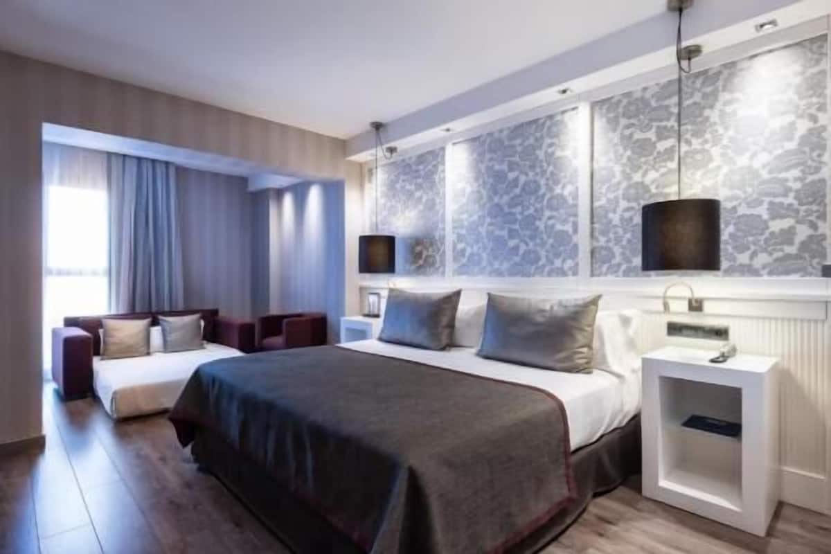 Ein modernes Hotelzimmer mit einem großen Bett, einer Sitzecke mit Sofa und Sesseln, einem Nachttisch, einer Lampe und Blick auf die Stadt durch das Fenster.