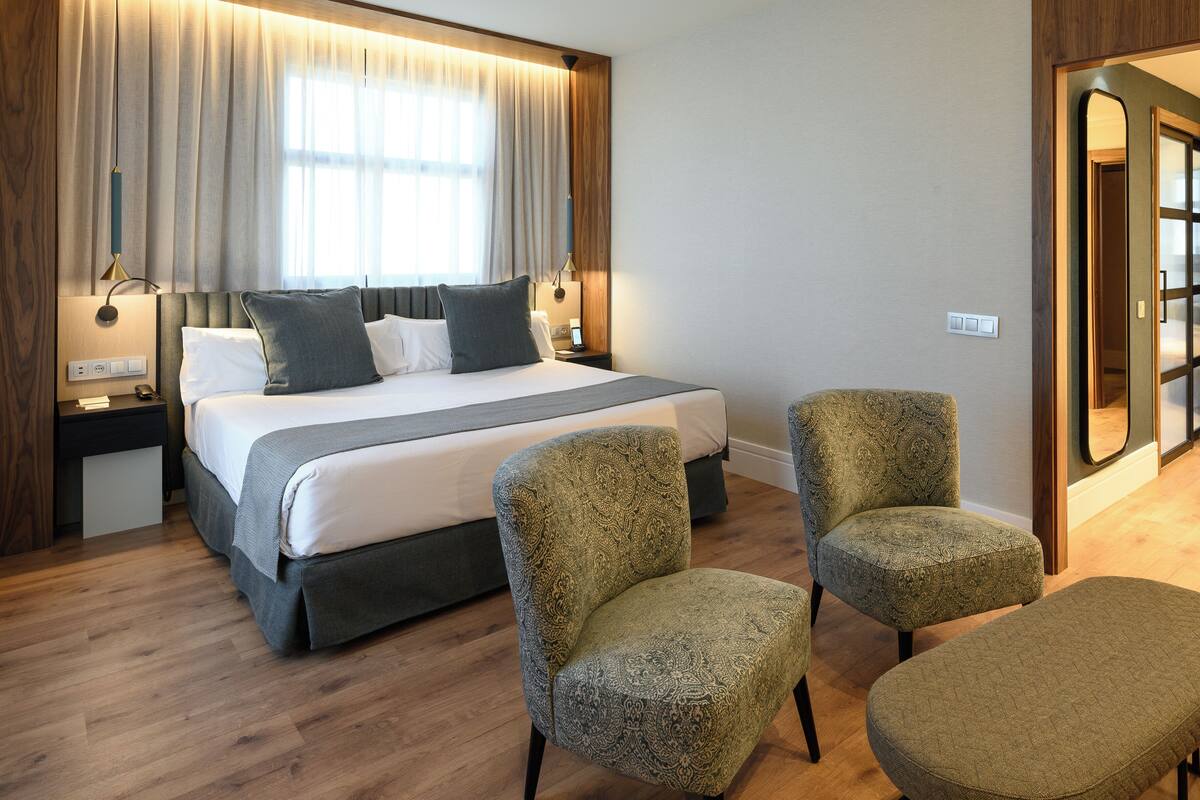 Une chambre d’hôtel avec un grand lit, deux fauteuils, une banquette, une table de chevet, une lampe et une fenêtre avec des rideaux.