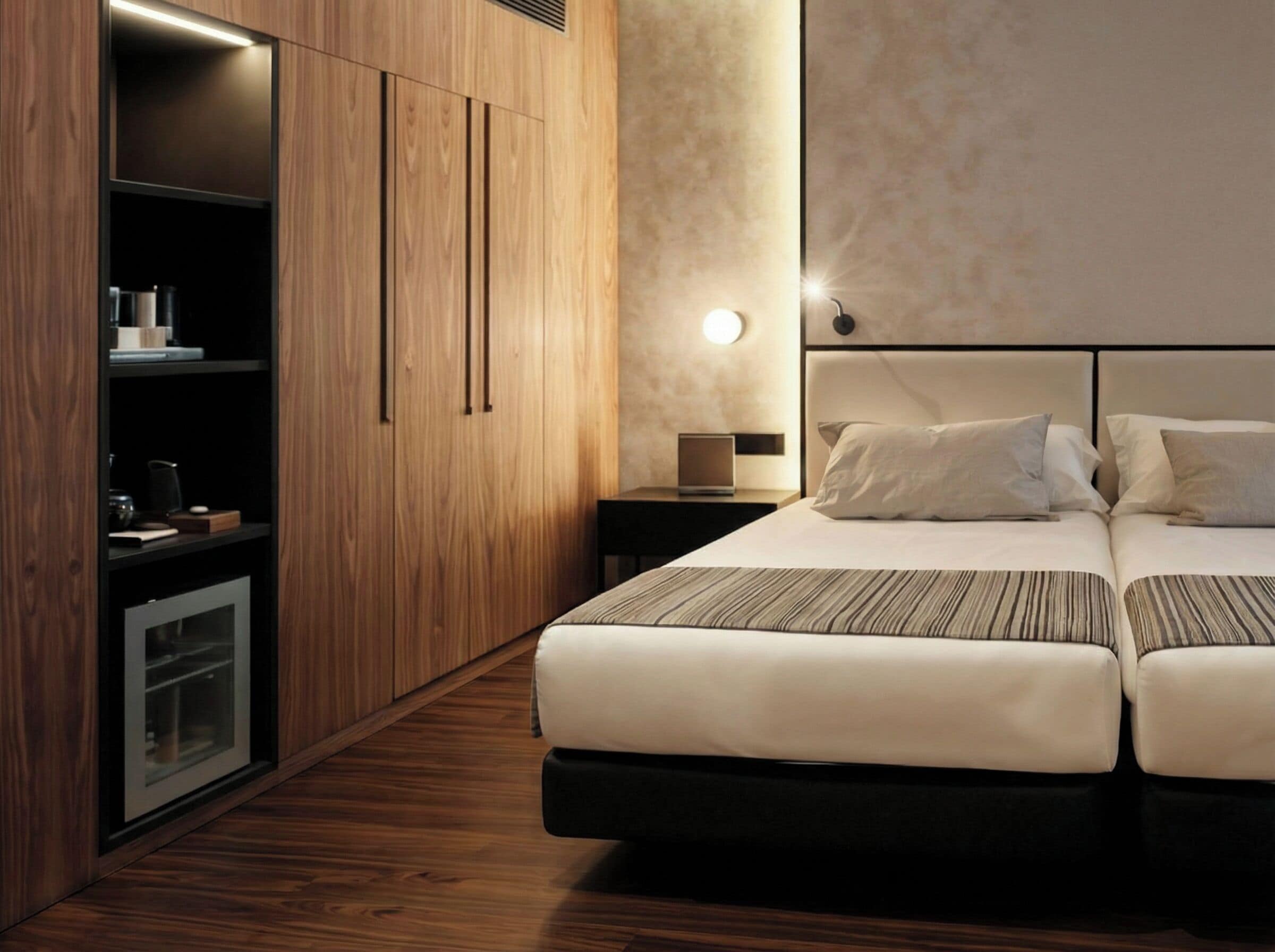 Habitación de hotel moderna con armario de madera, cama con dos almohadas, mesita de noche con lámpara y televisión integrada.