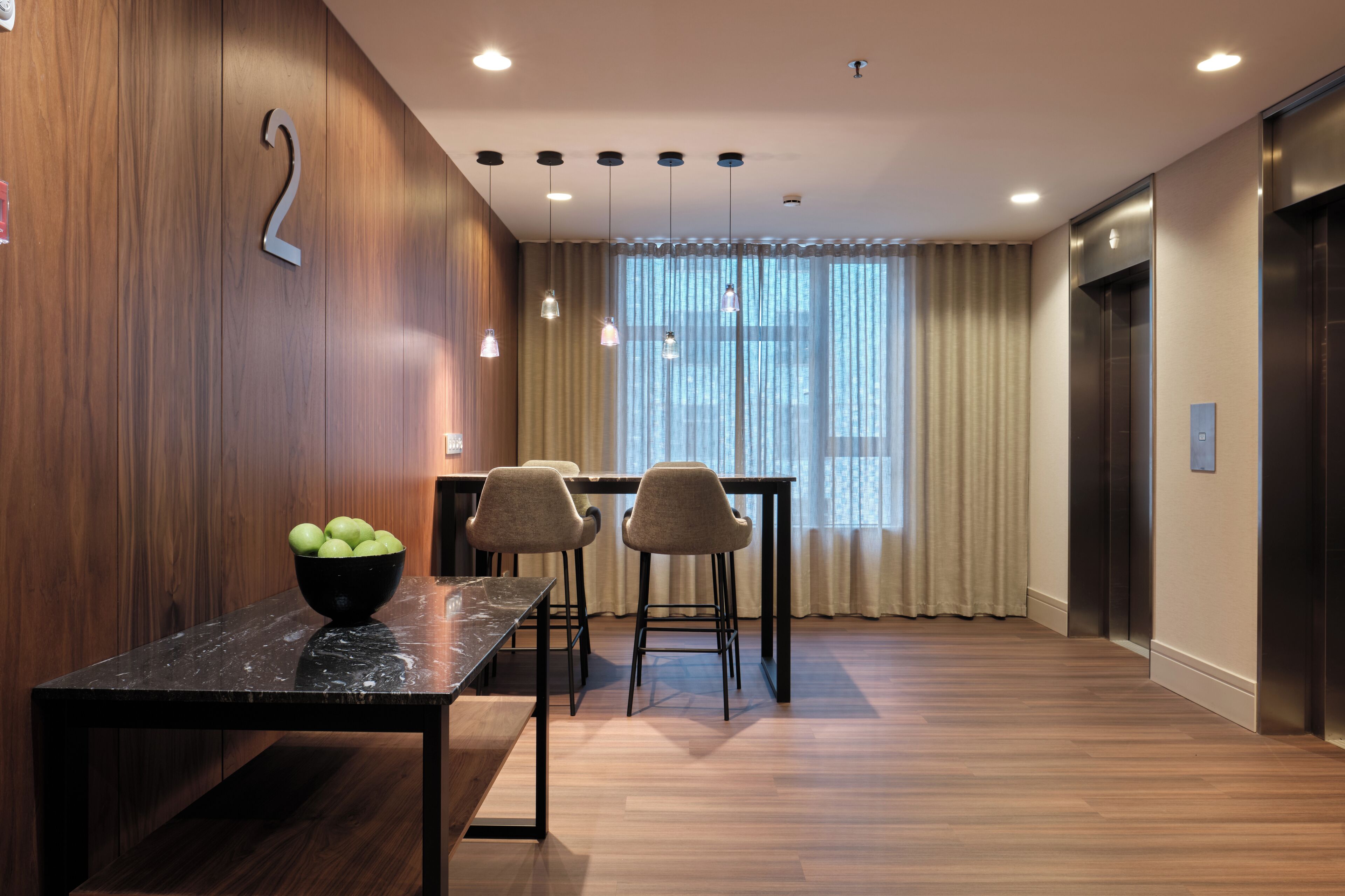 A modern hotel lobby with a wooden wall, a marble table with a bowl of apples, two bar stools, a window with curtains, and an elevator.