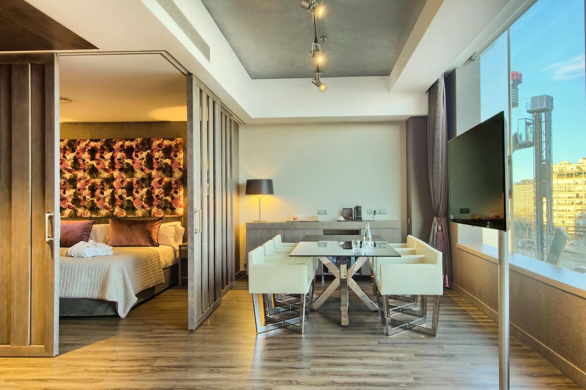 Une chambre d’hôtel moderne équipée d’un lit, d’un coin repas et d’une grande fenêtre donnant sur la ville.
