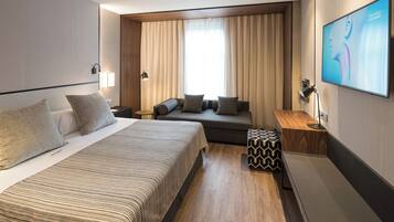 Una habitación de hotel moderna con una cama grande, un sofá, una mesita y un televisor fijado a la pared.