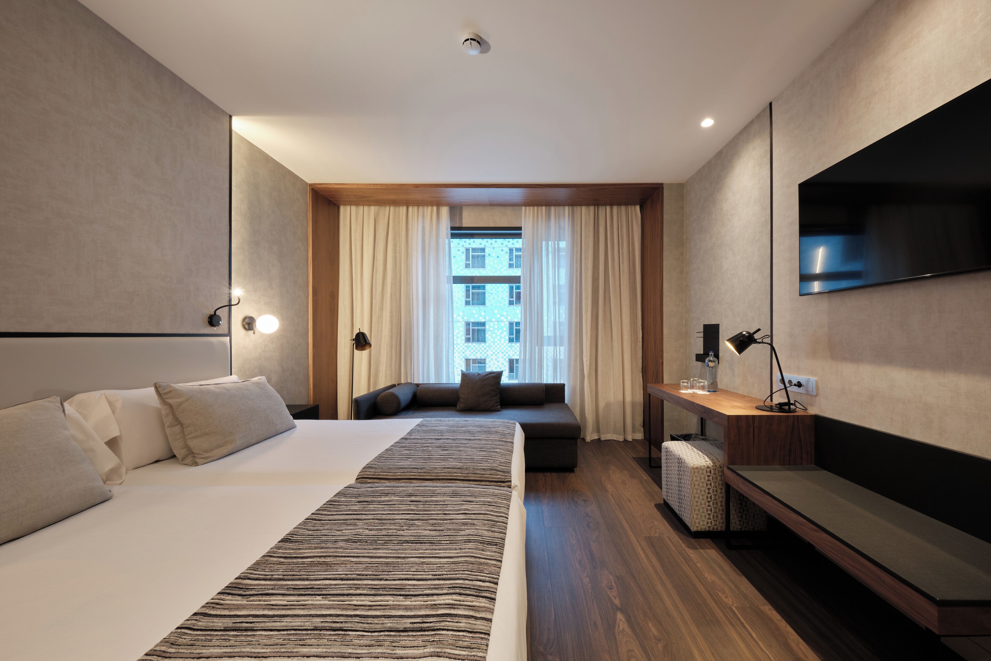 A modern hotel room with a large bed, a sofa, a desk, and a TV.