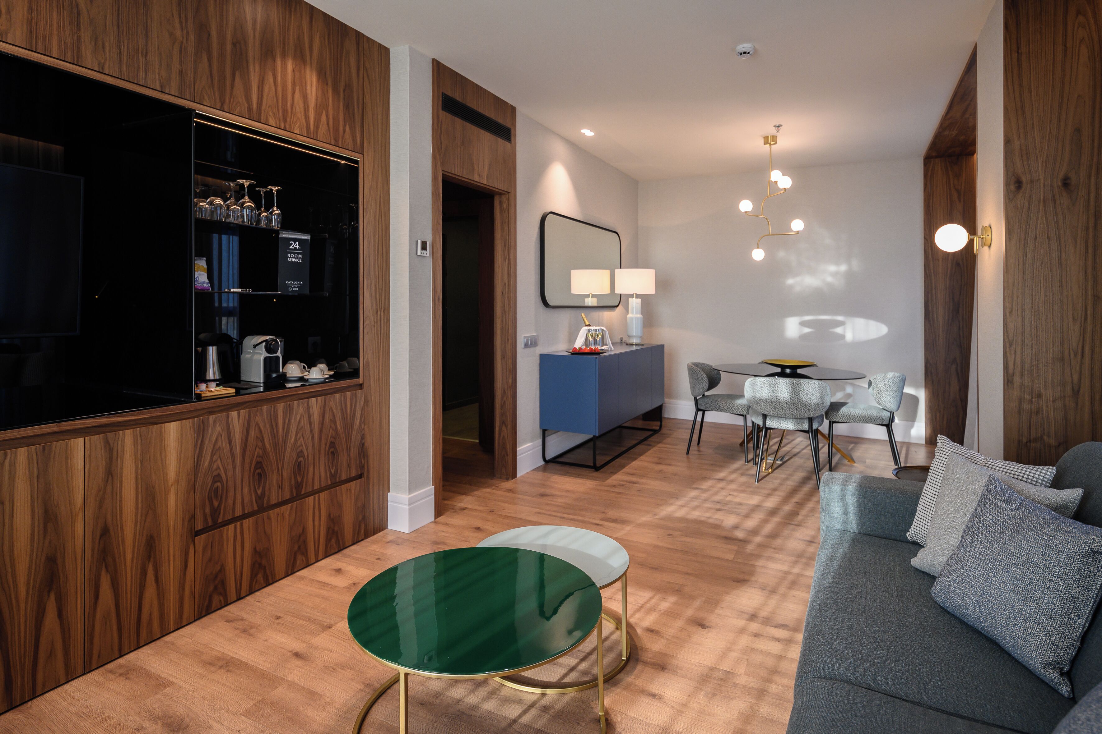 A modern living room with a sofa, a glass coffee table, a dining area with a table and chairs, and a kitchen area with a refrigerator and cabinets.