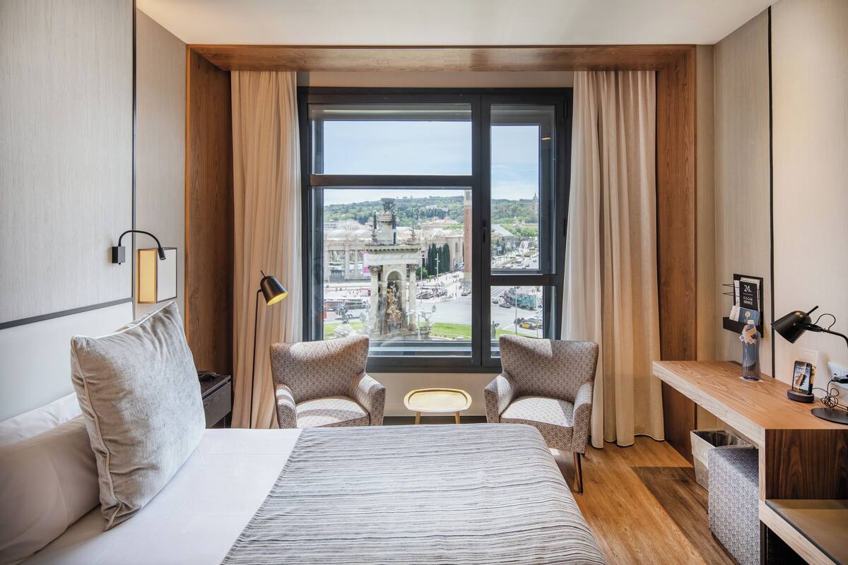 Une chambre d’hôtel avec un lit, deux fauteuils, un bureau, une vue sur la ville et une fenêtre avec des rideaux.