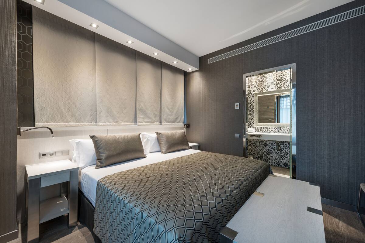 Une chambre d’hôtel moderne dotée d’un grand lit, d’une table de chevet, d’une banquette intégrée et d’un grand miroir.