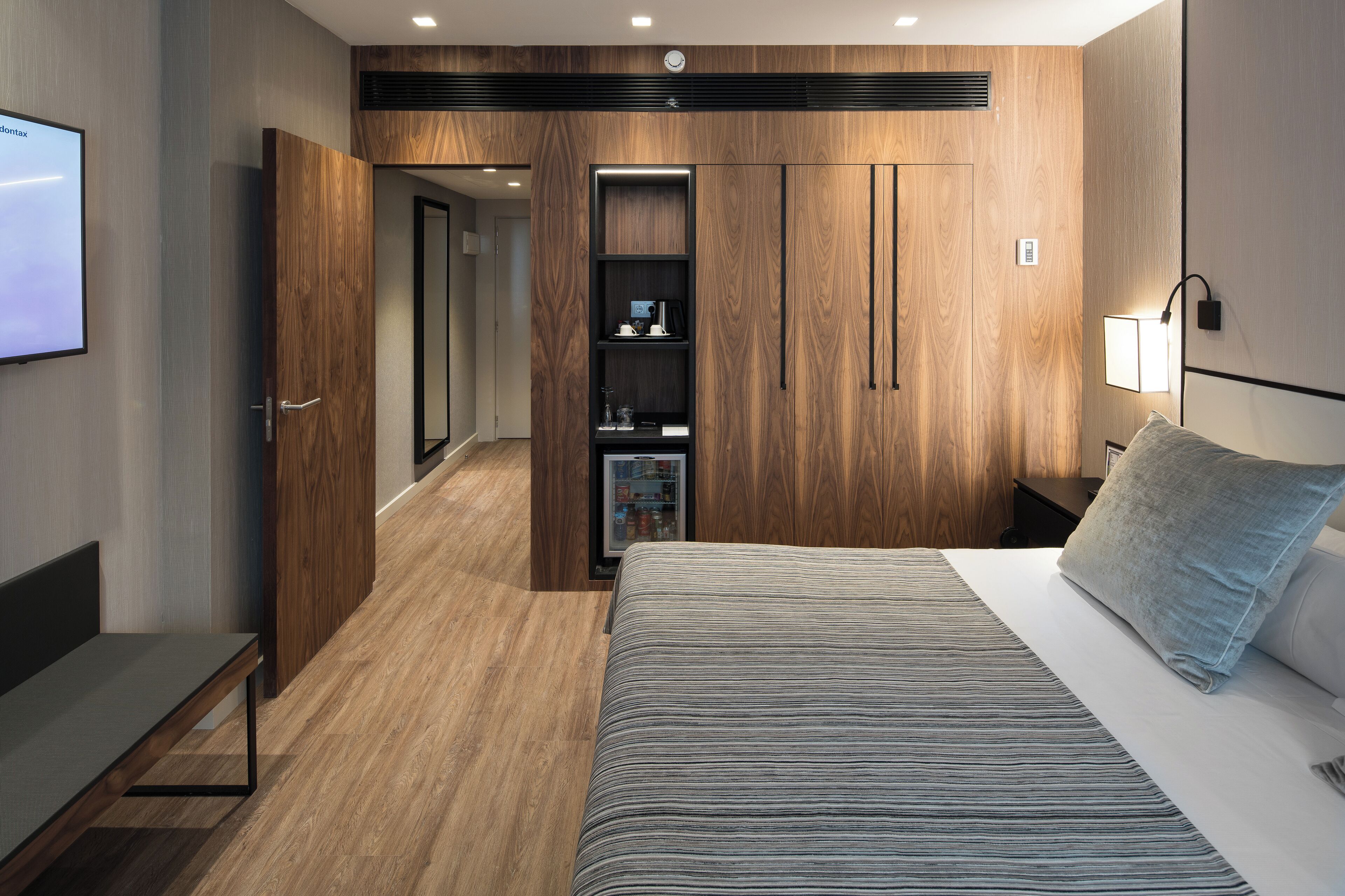A modern hotel room with a large bed, a built-in wardrobe, a bedside table, and a TV mounted on the wall.