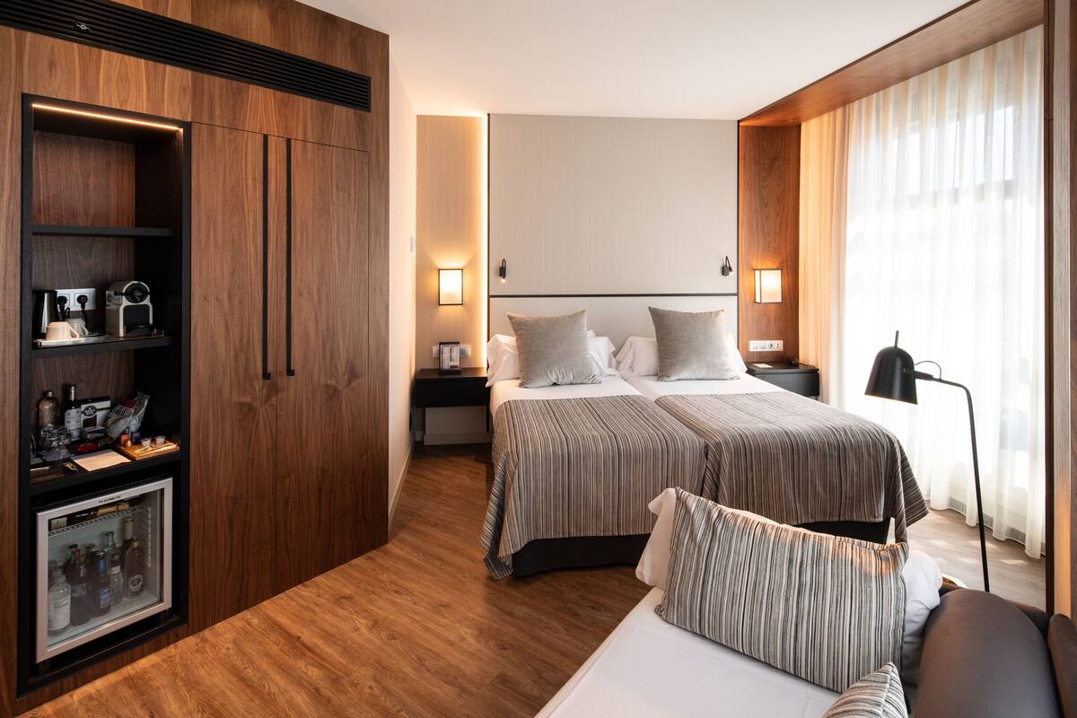 Une chambre d’hôtel moderne avec une armoire en bois, un minibar, un lit avec une literie à rayures et une table de chevet avec une lampe.