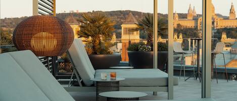A rooftop lounge with a view of a historic building, featuring a wicker chair, a sofa, and a small table with a drink.