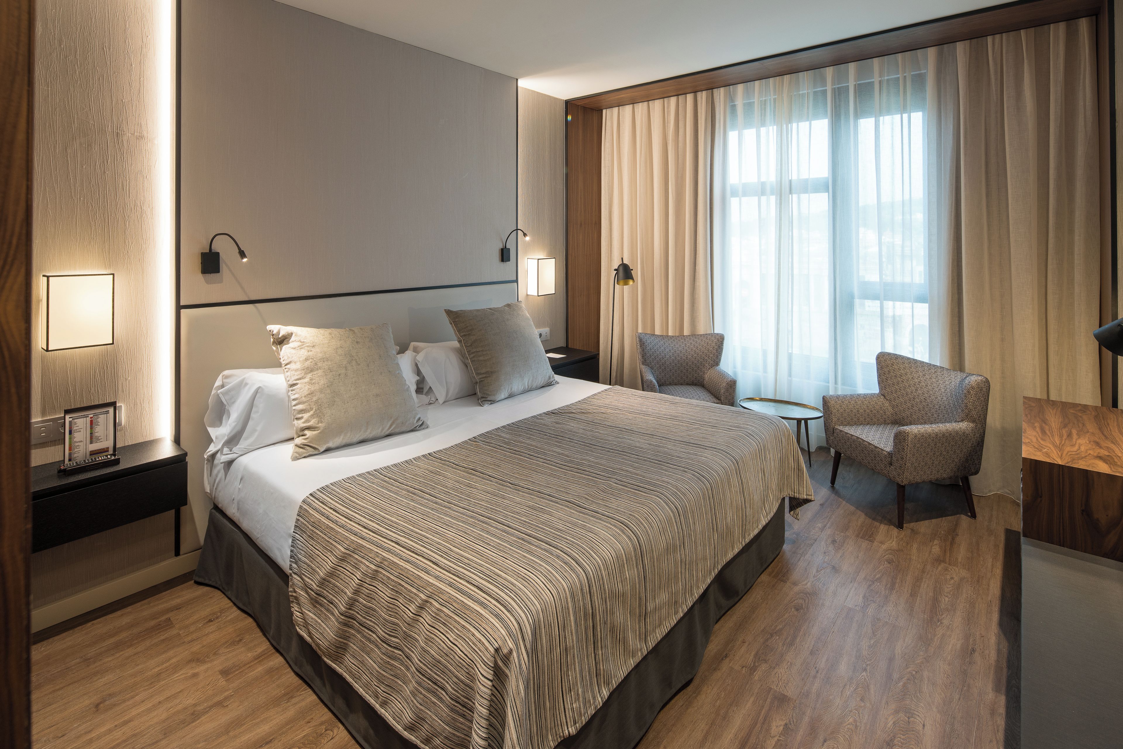 Quarto de hotel com uma cama grande, duas mesas de cabeceira, uma cadeira, uma escrivaninha e uma janela ampla com cortinas.