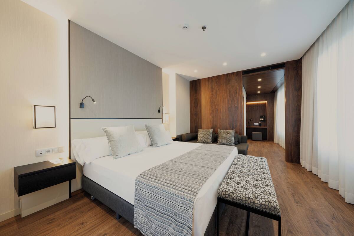 Une chambre d’hôtel moderne avec un grand lit, une table de chevet, un banc et une vue sur le couloir et la salle de bain.