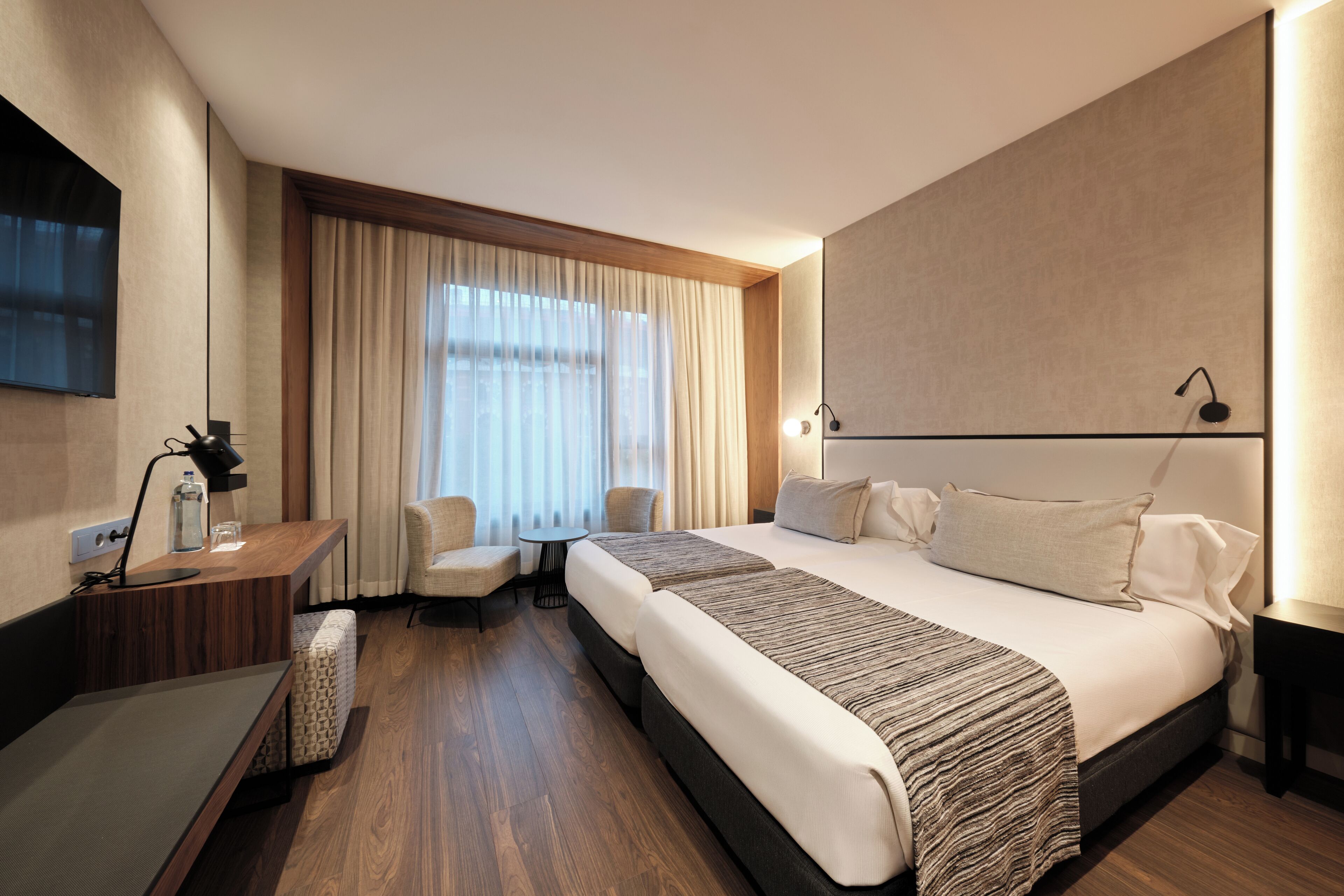 A modern hotel room with a large bed, a desk with a lamp, a chair, and a small table.