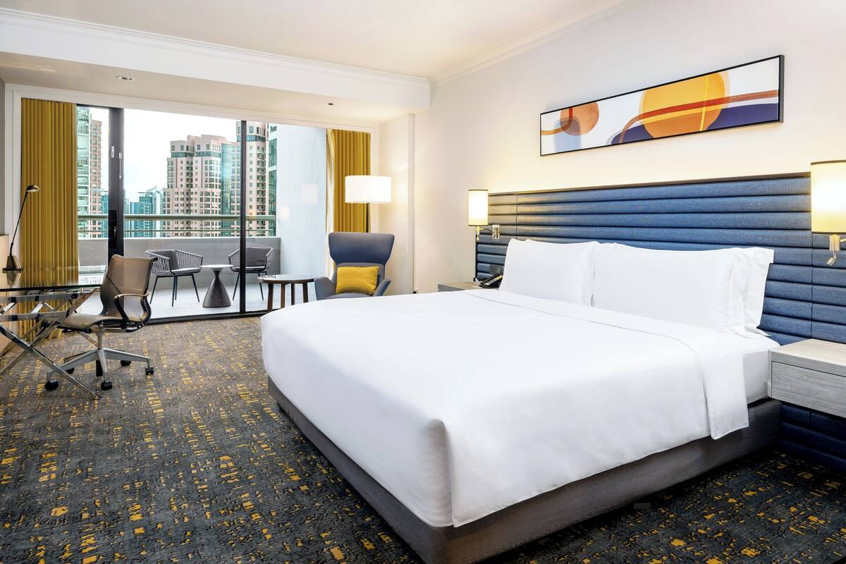 Habitación de hotel con una cama grande, un escritorio con silla, vistas a la ciudad a través de amplios ventanales y un cabecero moderno.
