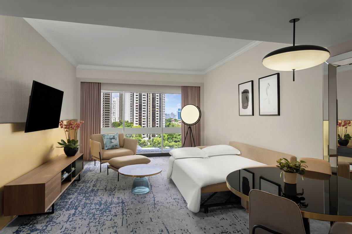 Una habitación de hotel moderna con sofá, cama, mesa de comedor, televisor y un gran ventanal con vistas a la ciudad.