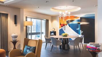 Quarto de hotel moderno com área de jantar, uma grande janela com cortinas, um candelabro e uma pintura abstrata colorida na parede.