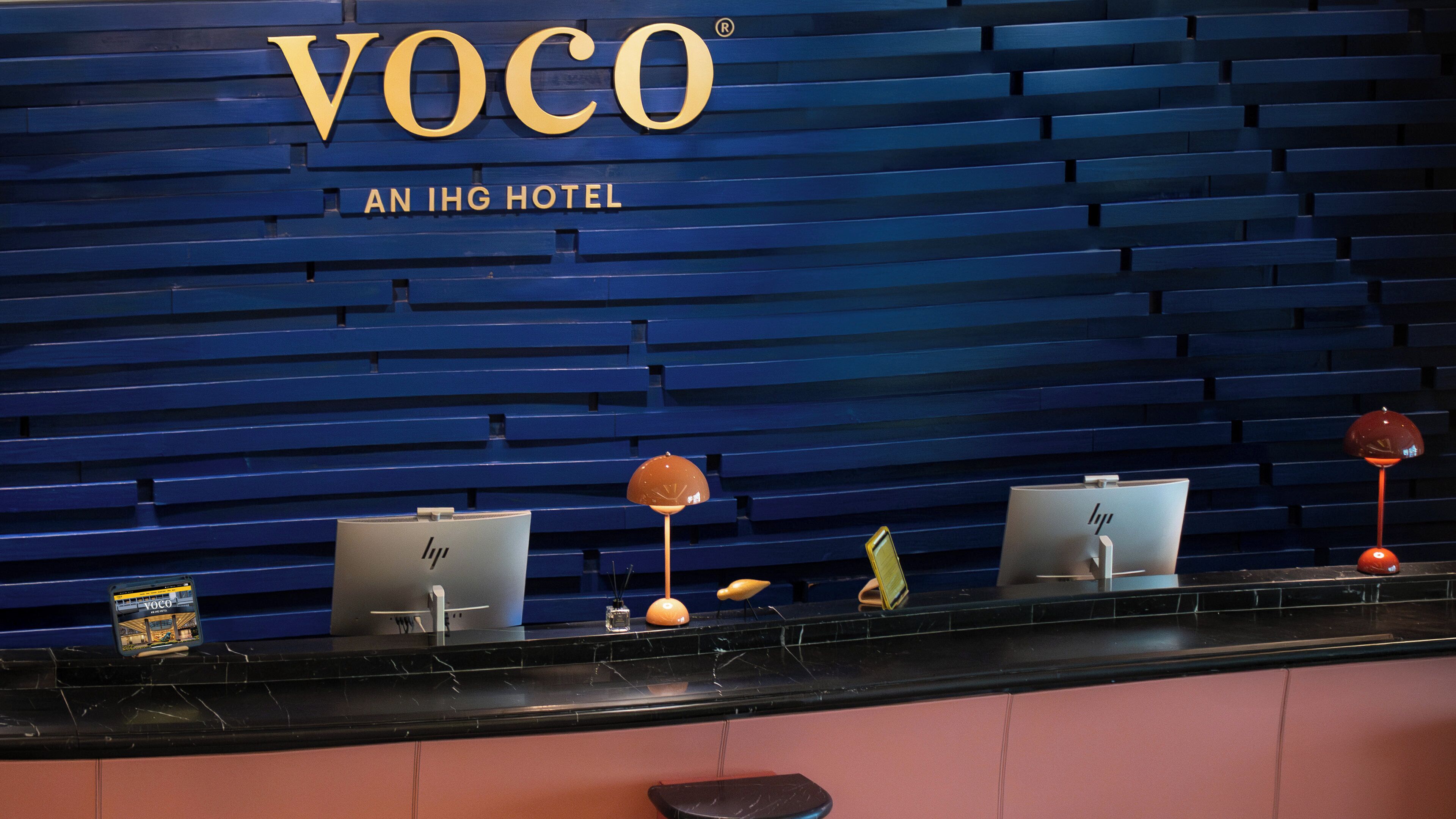 Una recepción con fondo de pared azul, un cartel que dice 'VOCO AN IHG HOTEL' y varios objetos, como un ordenador portátil, una lámpara y una caja pequeña.