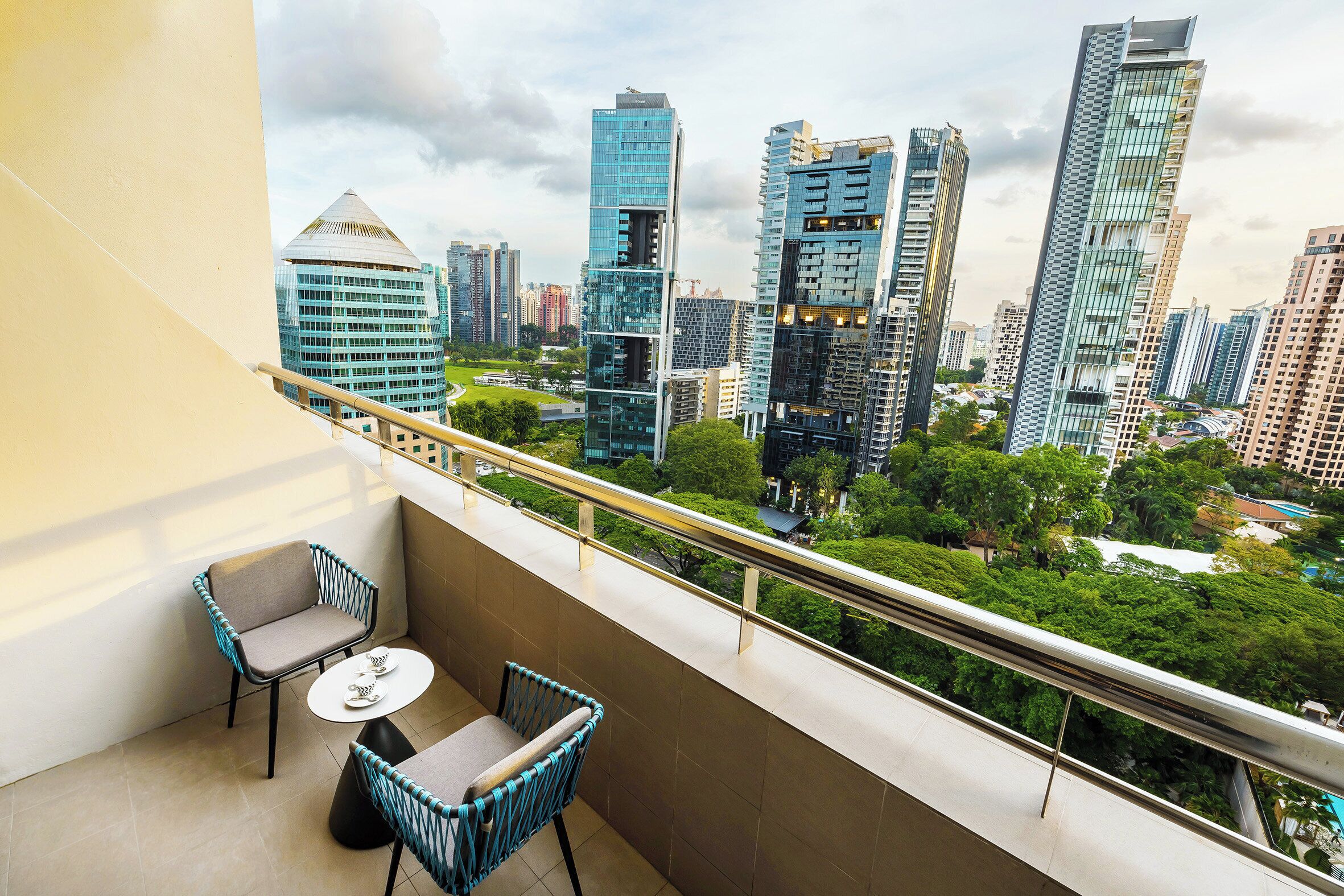 Un balcón con dos sillas y una mesa pequeña, con vistas a un paisaje urbano que incluye edificios modernos y áreas verdes.
