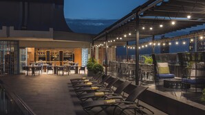 Un espace de restauration en plein air avec des tables et des chaises, un bar avec des étagères remplies de bouteilles, et une vue sur le paysage urbain au crépuscule.