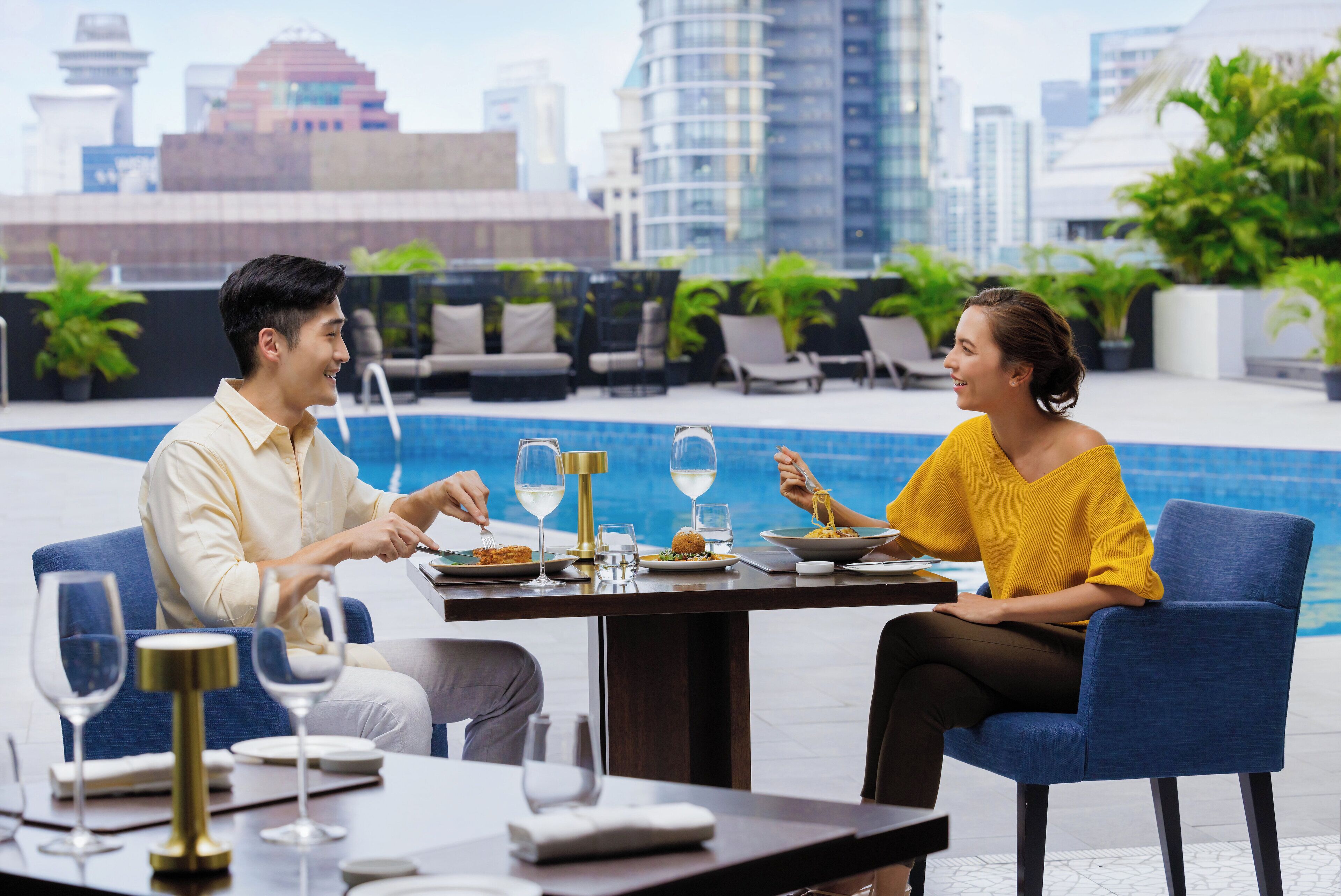 A dining table with plates, glasses, and cutlery, set against a backdrop of a city skyline and a pool.