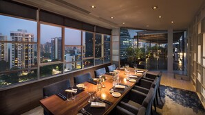 Un espace repas avec une longue table dressée pour le repas, de grandes fenêtres offrant une vue sur la ville et un aménagement intérieur moderne.