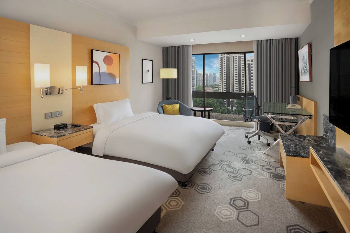 Habitación de hotel con dos camas, un escritorio, una silla y un amplio ventanal con vistas a la ciudad.