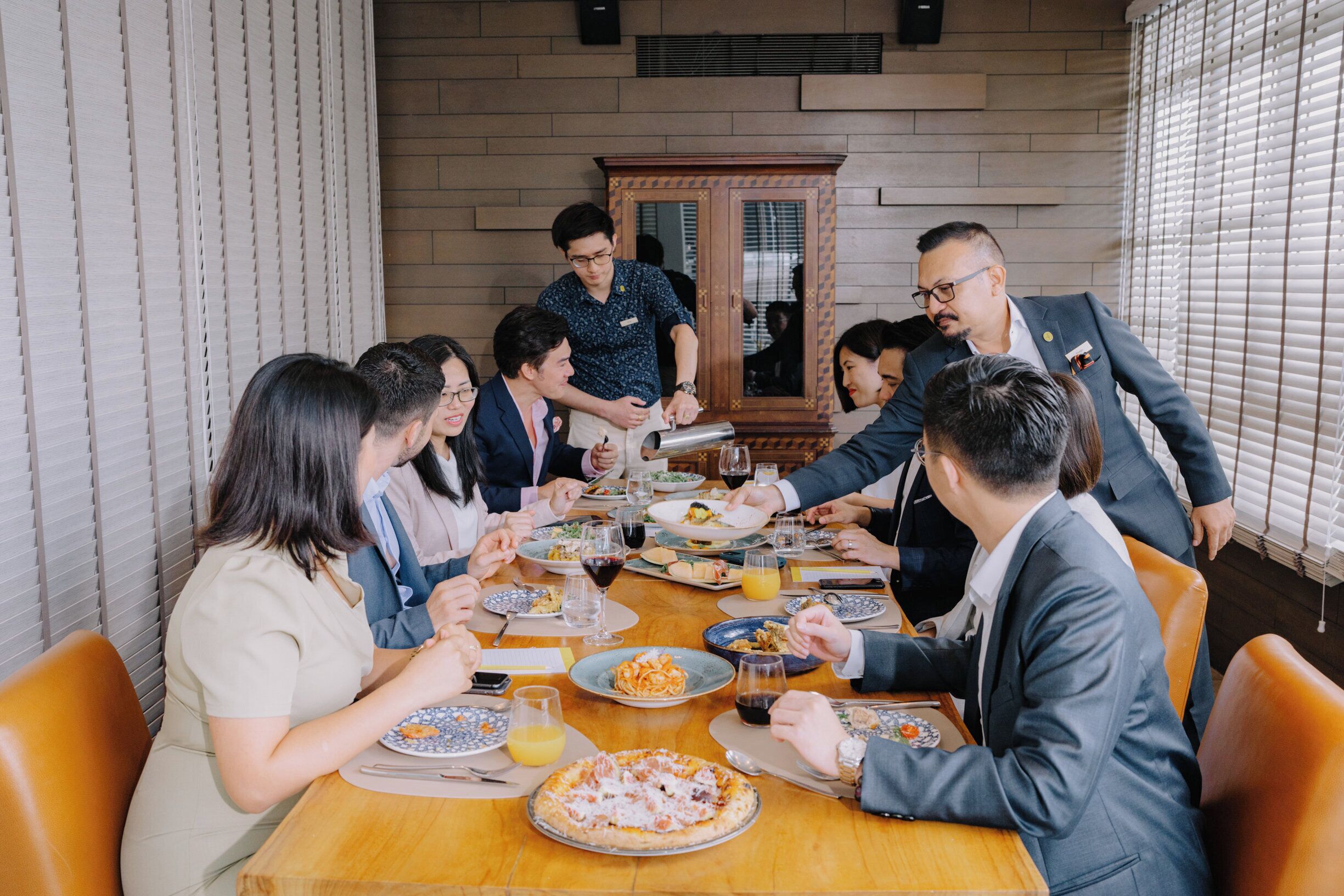 A group of people dining at a wooden table in a room with brick walls and a large mirror.