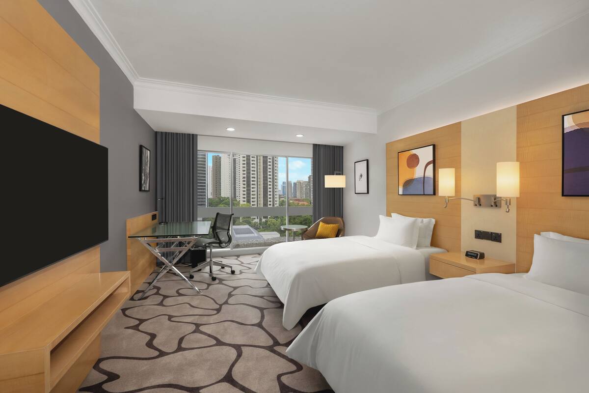 Habitación de hotel con dos camas, un escritorio, una silla, un televisor y un ventanal con vistas a la ciudad.
