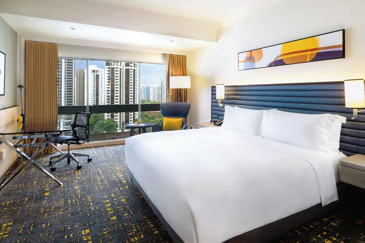 Habitación de hotel con una cama grande, un escritorio con silla, una ventana con vistas a edificios altos y un cabecero moderno.