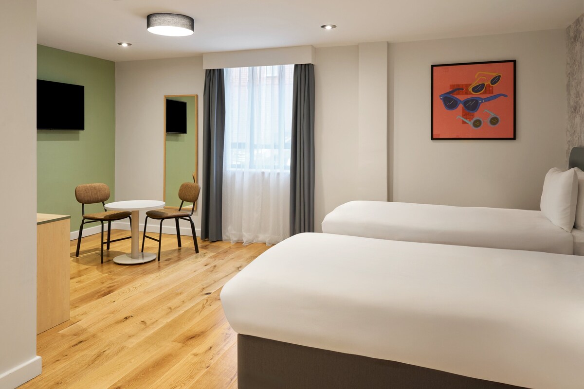 A hotel room with a wooden floor, a white bed, a small round table with two chairs, a TV mounted on the wall, and a window with curtains.