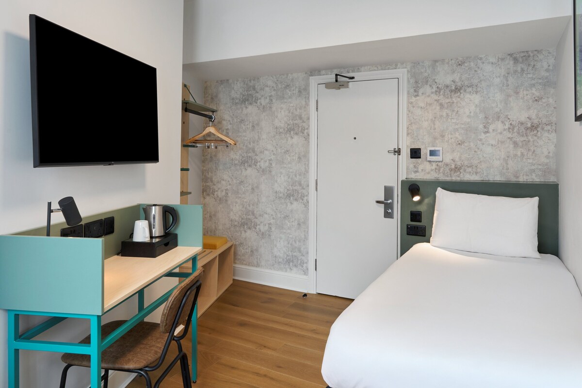 A hotel room with a bed, a desk with a chair, a television, and a door.
