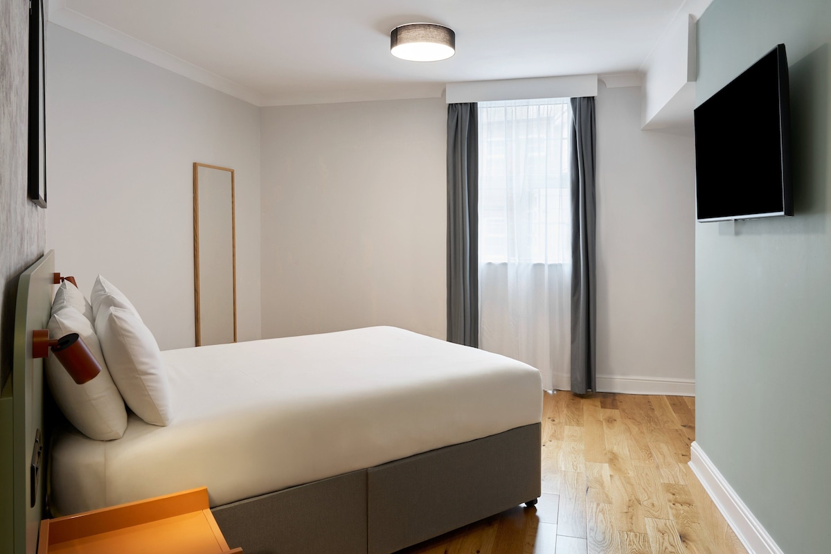 A modern hotel room with a large bed, a flat-screen TV mounted on the wall, and a window with curtains.