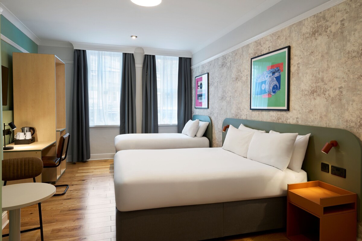 A hotel room with two beds, a desk, a chair, a small table, and a nightstand.