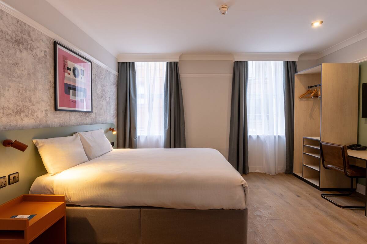 Hotellihuone, jossa on suuri sänky, puinen työpöytä, vaatekaappi ja seinällä oleva kehystetty taulu.