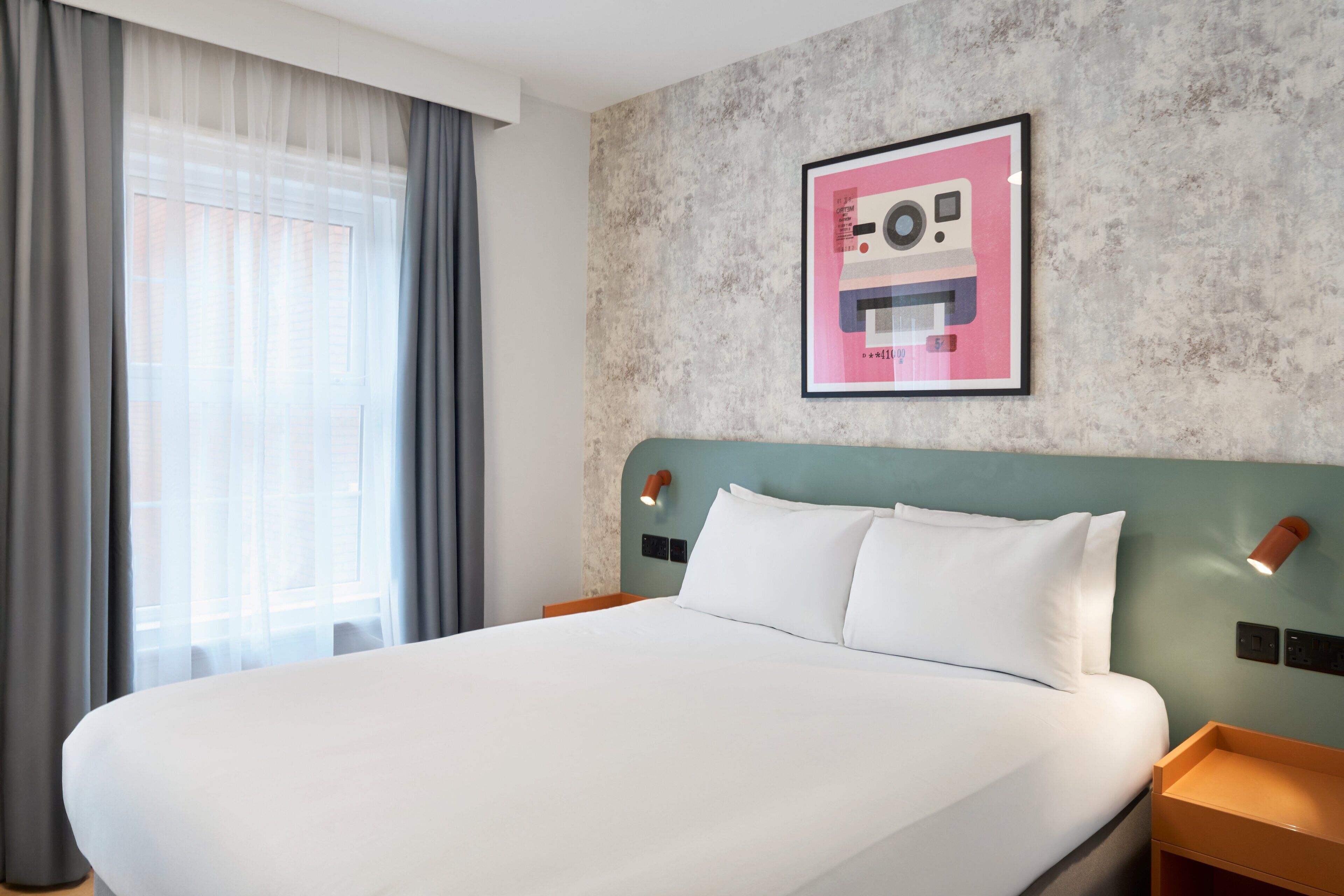 Una habitación de hotel con una cama grande, un cabecero con luces empotradas en la pared, un cuadro enmarcado y una ventana con cortinas.