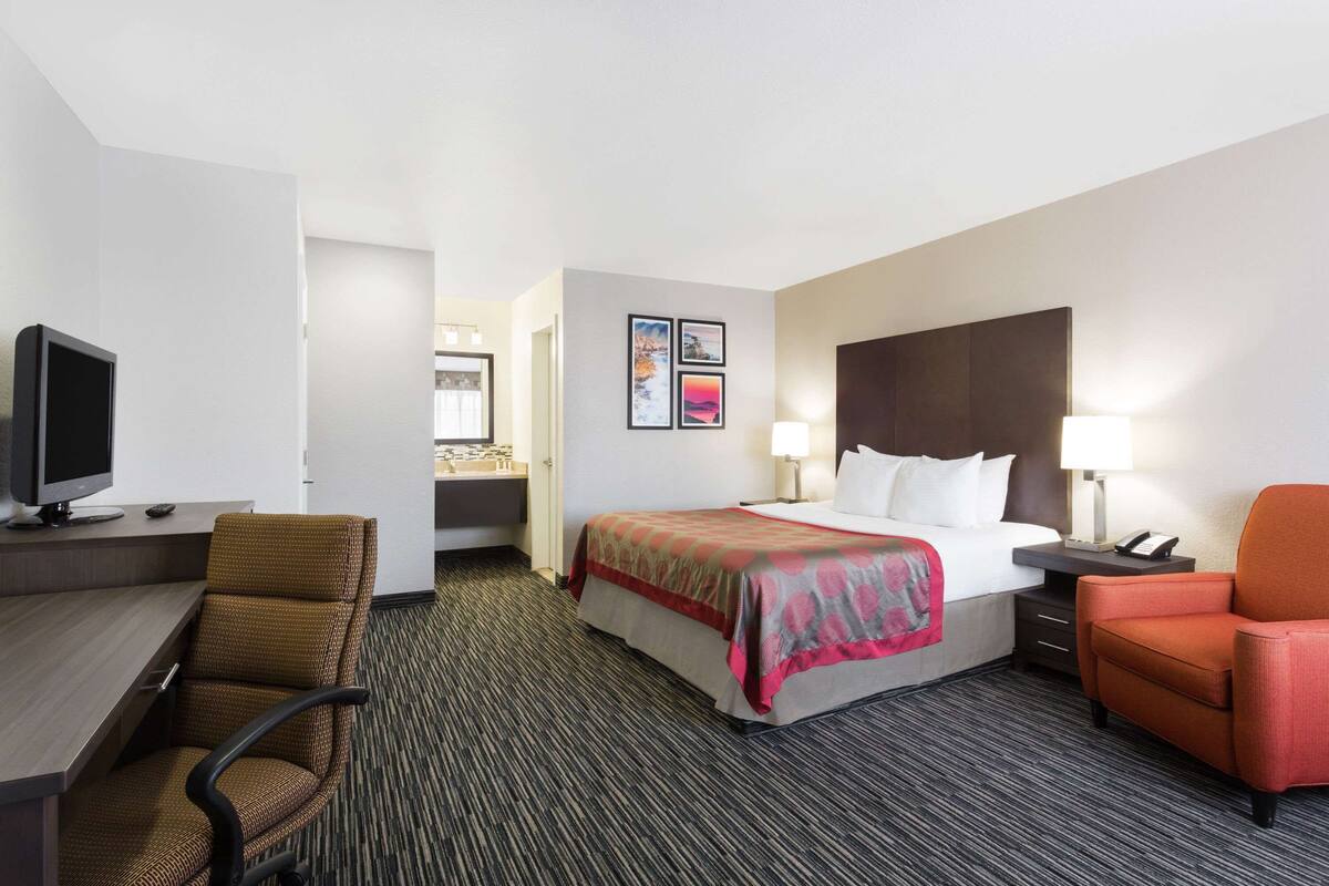 A hotel room with a bed, desk, chair, TV, and artwork on the wall.