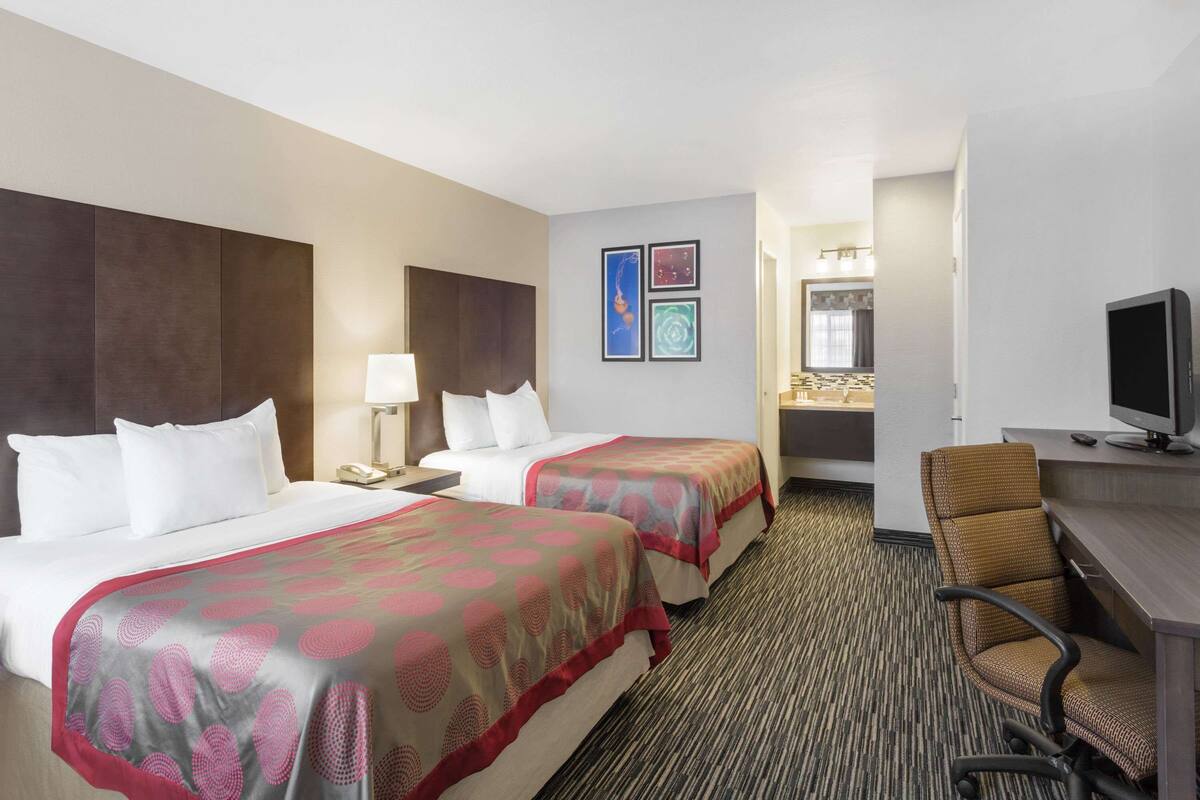 A hotel room with two beds, a desk with a chair, a TV, and a bathroom visible in the background.