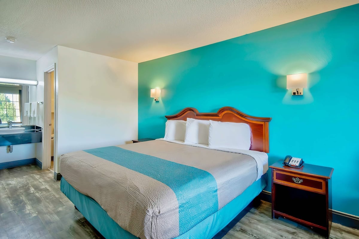 Une chambre d’hôtel avec un lit, une table de chevet, une lampe fixée au mur et une salle de bain visible à travers une porte ouverte.