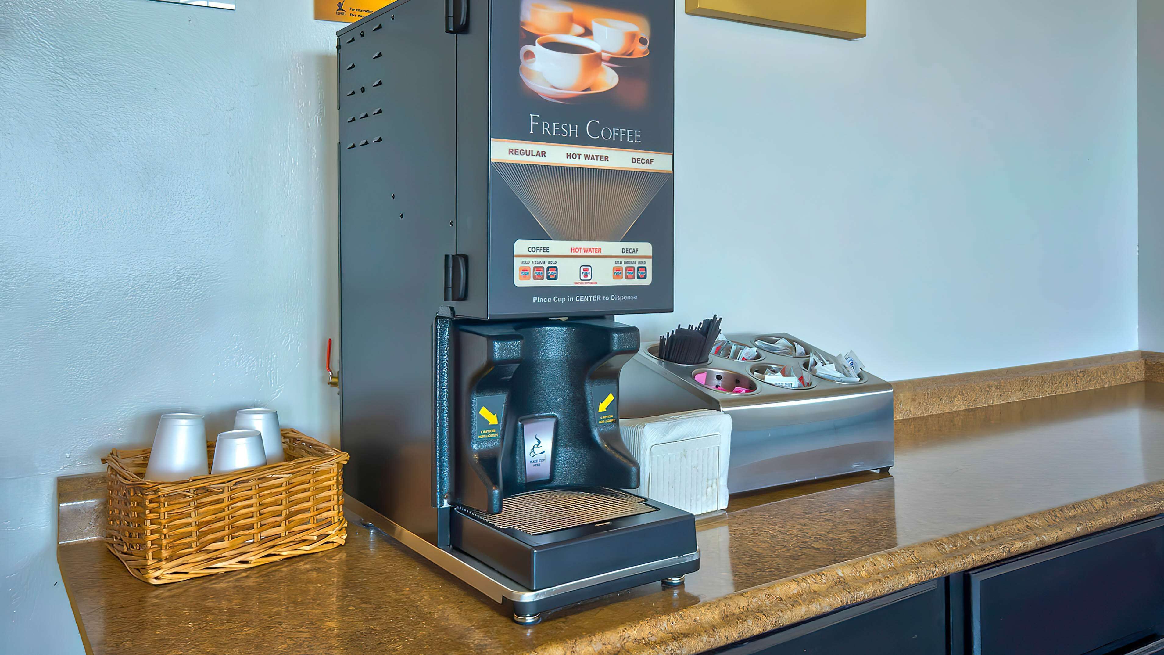 A coffee machine with a 'Fresh Coffee' sign, a basket of disposable cups, and a tray of condiments.