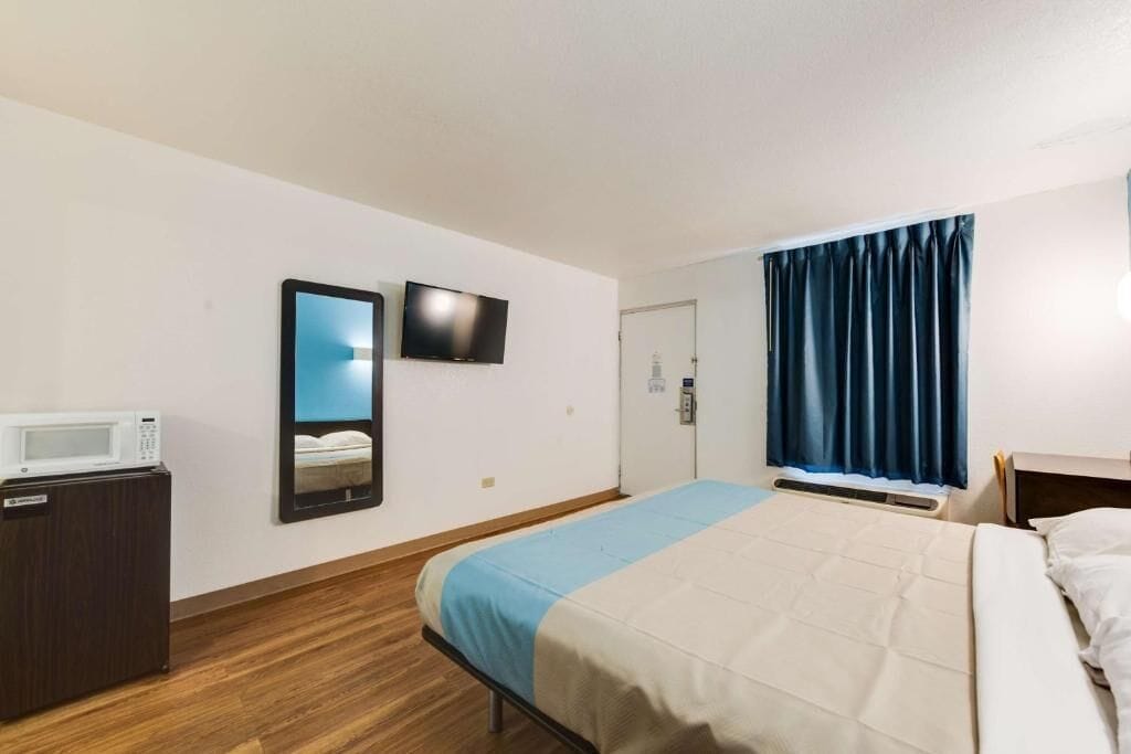 A hotel room with a bed, a TV mounted on the wall, a microwave on a wooden cabinet, and a window with dark curtains.