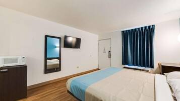 Quarto de hotel com cama, televisão presa à parede, micro-ondas sobre um armário de madeira e janela com cortinas escuras.