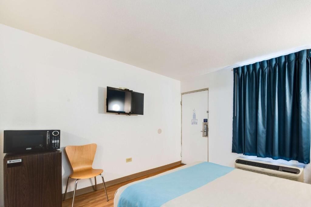 A hotel room with a bed, a television mounted on the wall, a desk with a computer, a chair, and a window with curtains.