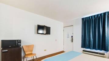 A hotel room with a bed, a television mounted on the wall, a desk with a computer, a chair, and a window with curtains.