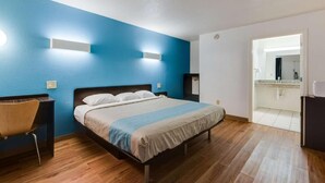 Un dormitorio con cama, una silla y un escritorio. Se ve un baño a través de una puerta abierta.