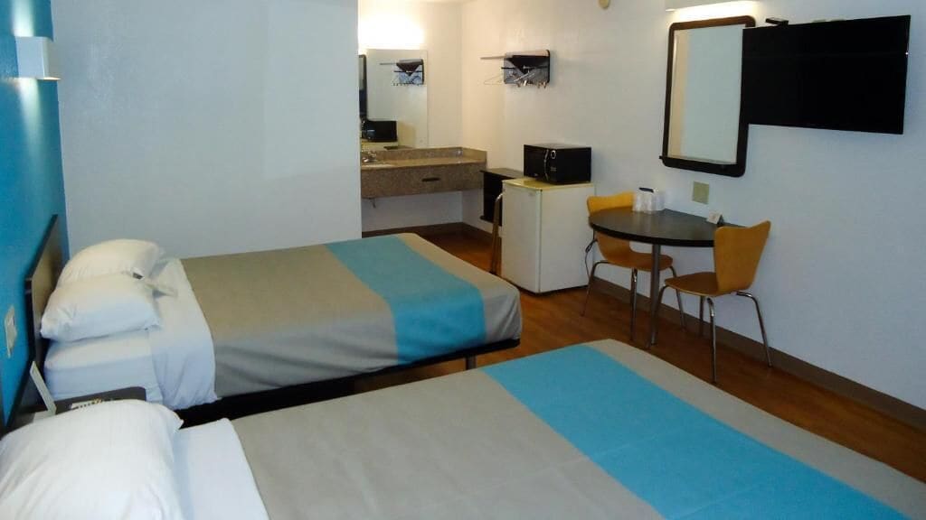 A hotel room with two beds, a desk, a chair, a small refrigerator, and a mirror on the wall.