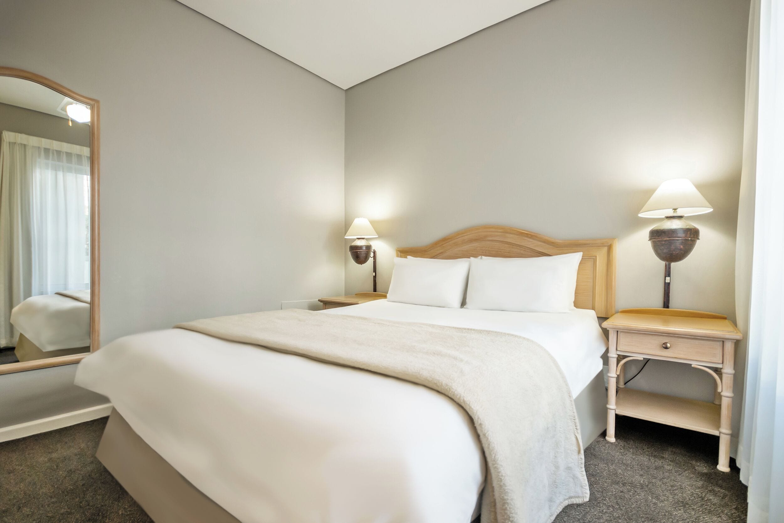 A neatly made bed with a headboard, two bedside lamps, a nightstand, and a large mirror.