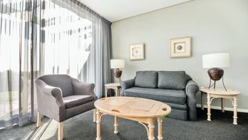 A modern hotel room with a grey sofa, a wooden coffee table, two side tables with lamps, and a large window with sheer curtains.