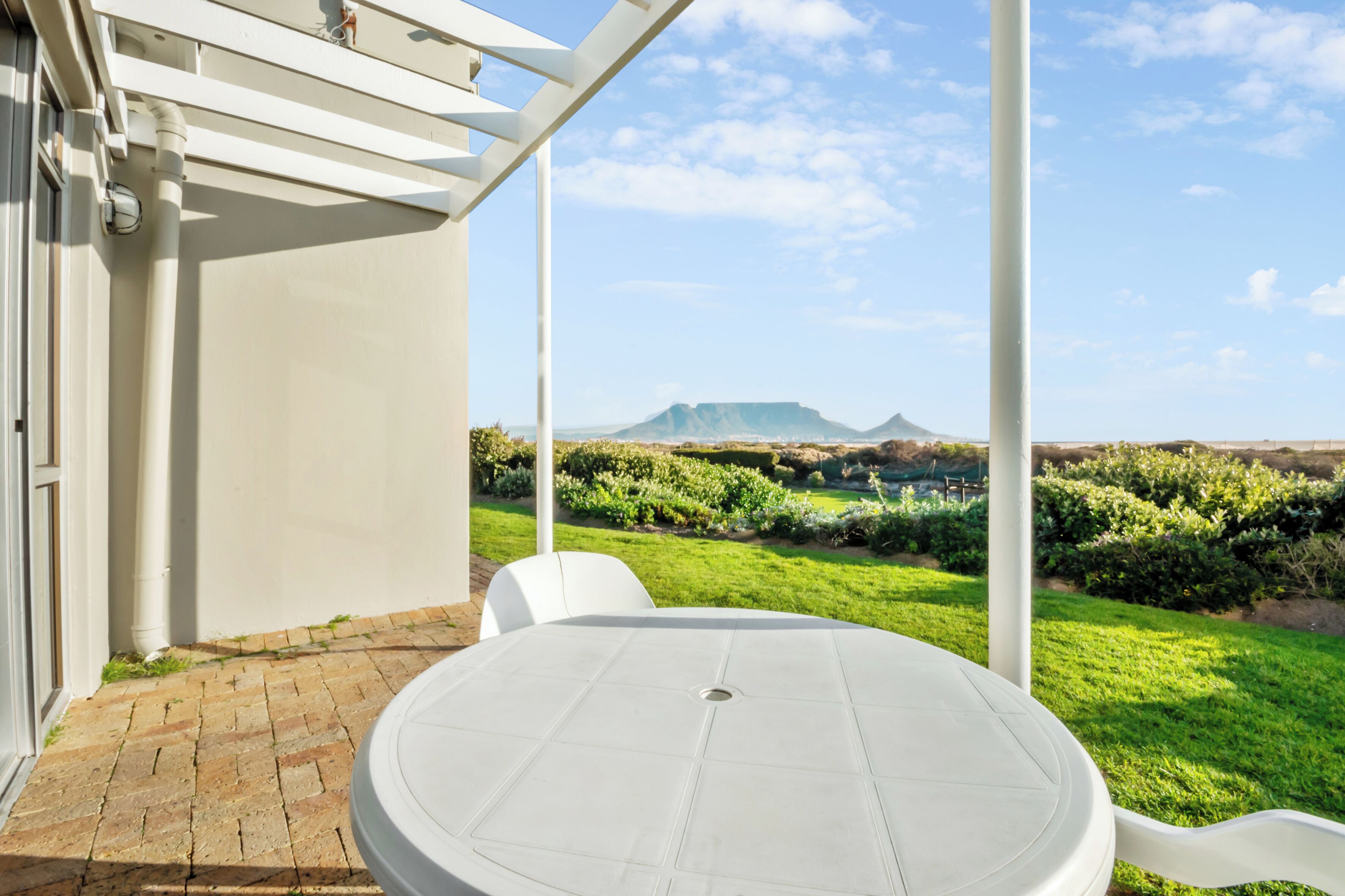 A patio with a round table and chairs, a white pergola, and a view of a green landscape with distant mountains.