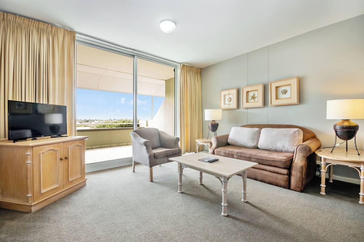 Ein Hotelzimmer mit Fernseher, Sofa, Sessel, Couchtisch und Blick durch ein großes Fenster.